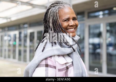 Smiling senior woman standing near modern entrance wearing pastel plaid coat gray scarf long dreads Stock Photo