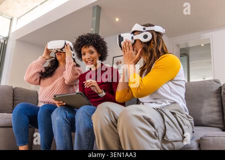 Three female friends using white VR headsets with center woman holding tablet on sofa at home Stock Photo