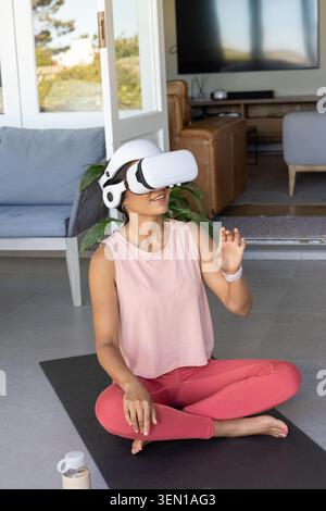 Woman sitting on black-mat at home in white VR-headset, pink-leggings and water-bottle nearby Stock Photo
