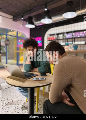 Two Young Men Collaborating Over Laptop and Phone in Casual Coworking Coffee Shop Meeting Stock Photo