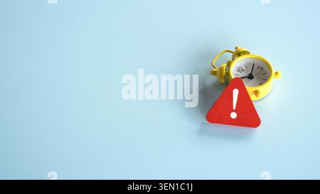 A small yellow vintage style alarm clock placed next to a red triangular warning sign with an exclamation mark. Stock Photo