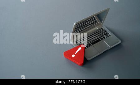 Miniature laptop computer on a grey background with a red warning exclamation mark icon sign representing cyber security alert and internet data dange Stock Photo