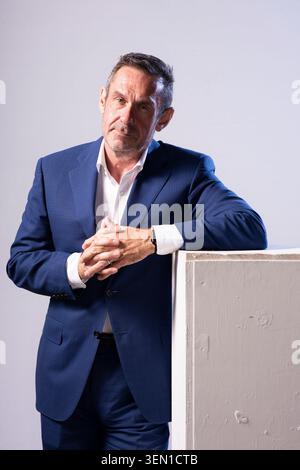 Paul Mason poses in a studio with a focused expression. His hands are clasped as he leans slightly on a white panel. The atmosphere shows a creative e Stock Photo