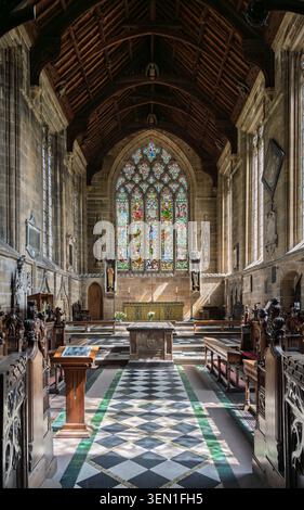 Symmetrical interior view looking down the chancel towards the high altar and large stained glass window with checkered stone floor tiling at Tideswel Stock Photo