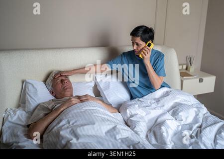 Worried elderly woman calling ambulance on smartphone while checking temperature of unwell husband Stock Photo