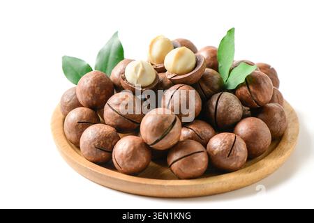 Premium macadamia nuts with cracked shells and creamy kernels arranged on a wooden plate with fresh green leaves. Isolated on a white background, this Stock Photo