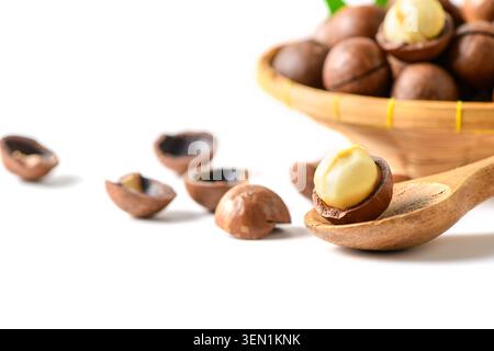 Close-up of a creamy macadamia nut kernel resting in its cracked shell on a wooden spoon, with scattered shells and a basket of whole macadamia nuts s Stock Photo