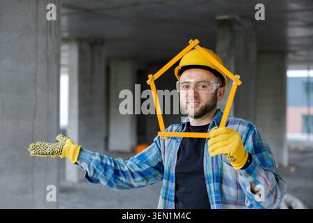 Smiling construction worker holding a house shape ruler, conceptualizing new home-building plans inside an unfinished building Stock Photo