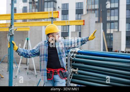 Construction worker in hard hat and safety glasses supervising building site development, extending arm measuring or indicating Stock Photo
