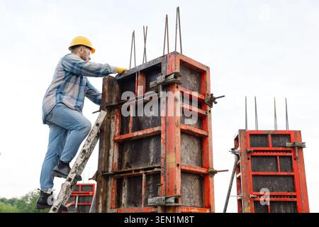 Construction worker in hard hat and safety glasses climbing ladder, setting metal formwork for a reinforced concrete column Stock Photo