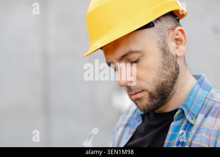 Thoughtful construction worker wearing a yellow hard hat and plaid shirt, concentrating on work at a building site Stock Photo