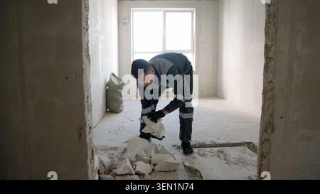 Male builder in protective uniform clearing concrete rubble inside unfinished room during home renovation project. Natural light from window highlight Stock Photo