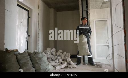 Male builder in workwear holds heavy sack amidst rubble and concrete blocks during renovation. Dusty interior with exposed walls and plastic covered w Stock Photo
