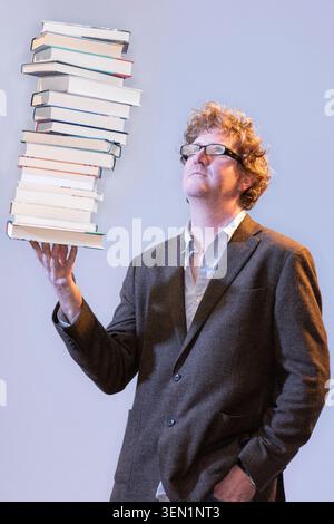 Shaun Bythell stands in a studio holding a tall stack of books on one hand. He looks upward with a thoughtful expression while wearing a brown suit an Stock Photo