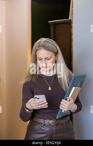 Business woman holding laptop and checking smartphone while standing in modern office environment. Concept of multitasking, communication, remote work Stock Photo