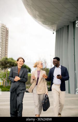 Three business people walk down a pathway in a city. They talk and smile while passing modern buildings. The weather is cloudy, and they are dressed i Stock Photo