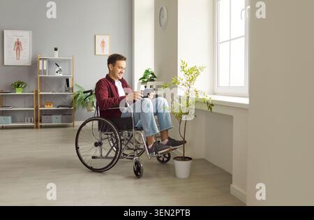 Happy young man in wheelchair using digital tablet, enjoying online communication and entertainment. Stock Photo