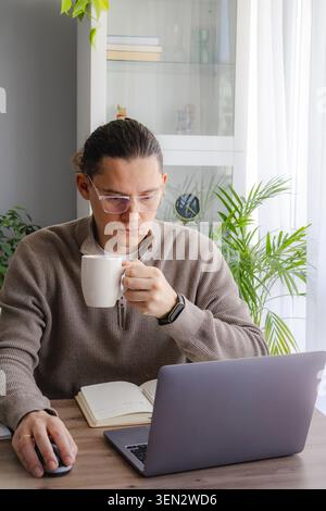 Middle eged man working remotely from home with laptop while drinking coffee at modern desk. Productive freelance morning routine, online business man Stock Photo