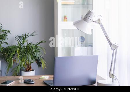 Modern workspace with laptop, smartphone and desk lamp on wooden table in bright home office interior. Minimal business background with copy space, co Stock Photo