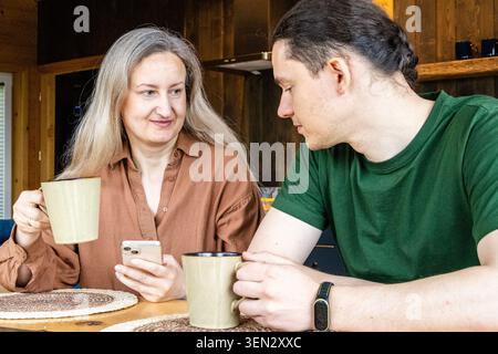 Couple drinking coffee and using smartphone together at the table in cozy mountain cabin in Zakopane, Poland. Relaxed travel lifestyle with rustic int Stock Photo