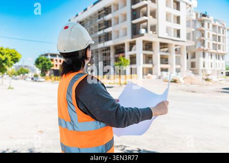 woman wearing a hard hat and safety vest inspects a construction site. She holds a clipboard and looks at a building under construction. Stock Photo