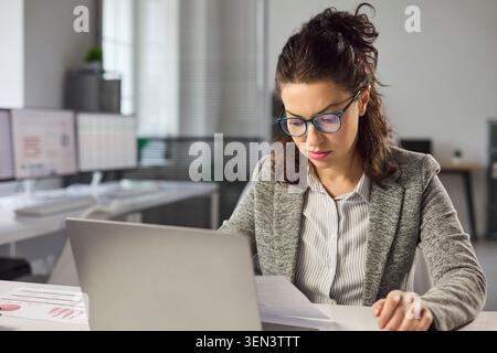 Focused female accountant analyzing financial report with laptop in modern office Stock Photo