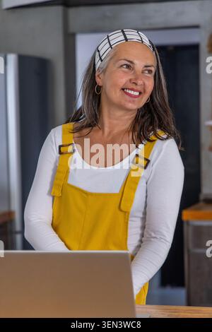 Senior woman wearing yellow overall dress standing behind counter in kitchen, using silver laptop Stock Photo