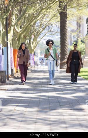 Three friends walking down tiled promenade under canopy, carrying coffee cup and crossbody bag Stock Photo