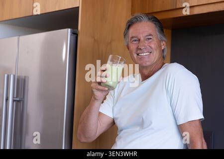 Senior man sitting on stool in home kitchen holding green smoothie glass and smiling at camera Stock Photo