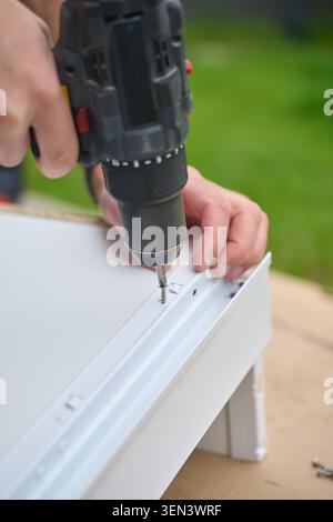 Man uses cordless drill to fasten screw in white furniture panel. Concept of DIY furniture assembly and home improvement Stock Photo