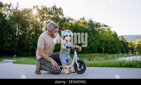Father teaching toddler son to ride balance bike while wearing safety helmet. Stock Photo