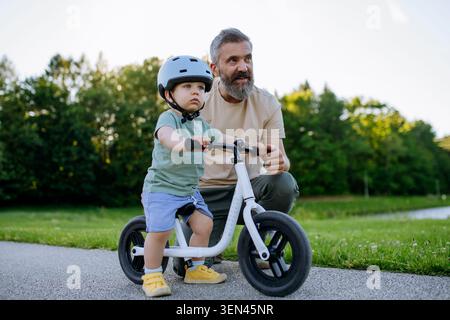 Father teaching toddler son to ride balance bike while wearing safety helmet. Stock Photo
