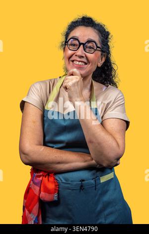 Mature woman wearing an apron and glasses, smiling with hand on chin, thinking Stock Photo