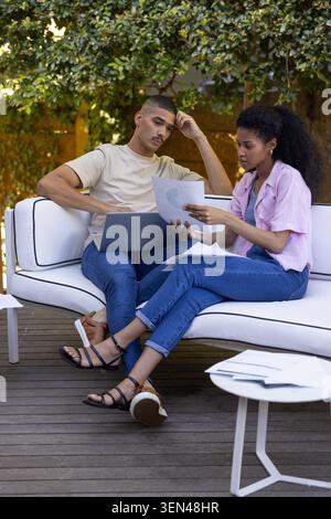 Diverse collaborators reviewing printed charts and typing on laptop on patio deck. Outdoor, terrace, greenery, teamwork, planning, documents, graphs Stock Photo