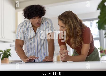 Couple leaning over white kitchen countertop using tablet, man in striped shirt, woman in rust top Stock Photo