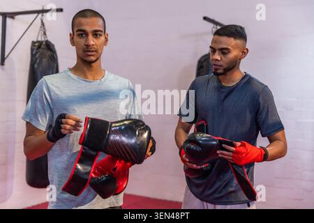 Two male boxing partners holding gloves and adjusting red hand wraps in t-shirts at boxing gym. Boxer, training, fitness, shorts, mats, brickwall, stu Stock Photo