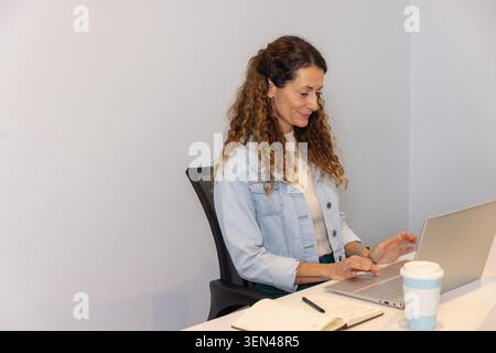 Woman sitting at desk in light-gray corner, working on laptop with notebook and mug, copy space Stock Photo