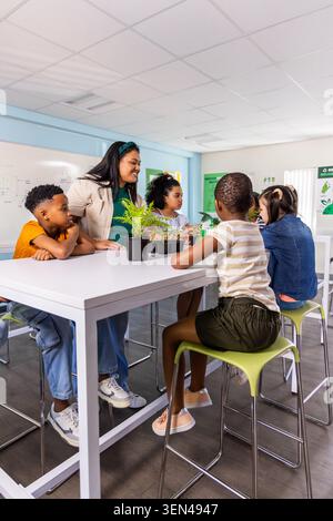 Teacher guiding four school-age children in classroom at white table, examining seedlings Stock Photo
