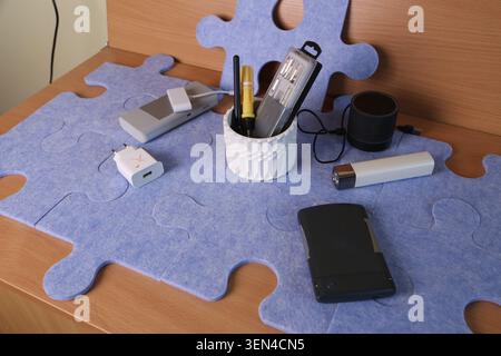 Several office supplies are placed on a blue puzzle mat in a workspace. The items include pens, a charger, and a flashlight. Natural light fills the room. Stock Photo