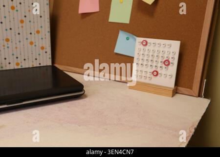 A desk is set up with a closed laptop on the left. A calendar sits on a stand, and colorful sticky notes are pinned to a corkboard in the background. Stock Photo