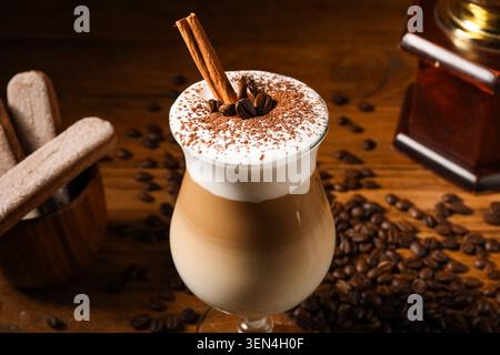 Layered latte macchiato with cinnamon stick, coffee beans, cocoa powder, and ladyfingers on rustic wooden table Stock Photo