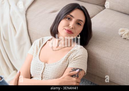 Relaxed young woman with short dark hair and gold jewelry leaning against a beige sofa in a cozy home setting, she wears a light-colored top and has a Stock Photo