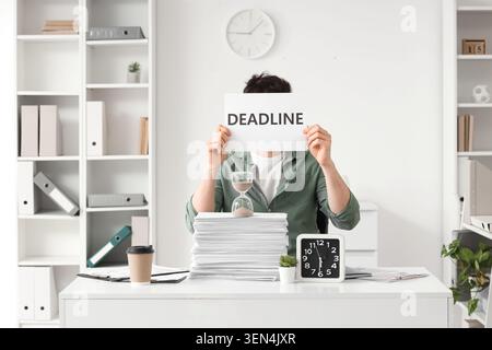 Overwhelmed office worker holding deadline sign with hourglass and stack of papers on desk, time management pressure Stock Photo