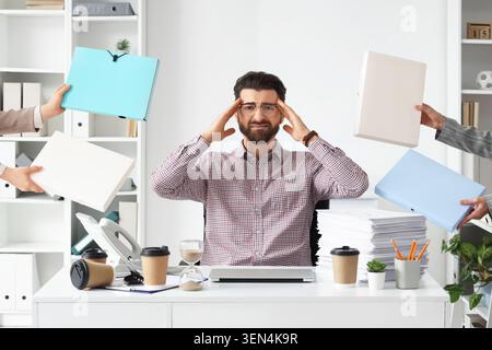 Overwhelmed businessman experiencing stress and headache from excessive workload and demanding tasks in office, hands from various directions present Stock Photo