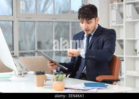 Shocked young businessman checking wristwatch for urgent deadline in modern office environment, a young businessman with a shocked expression checks h Stock Photo