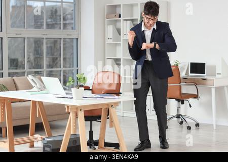 Excited young businessman checking smartwatch in modern office, celebrating success and timely achievement Stock Photo