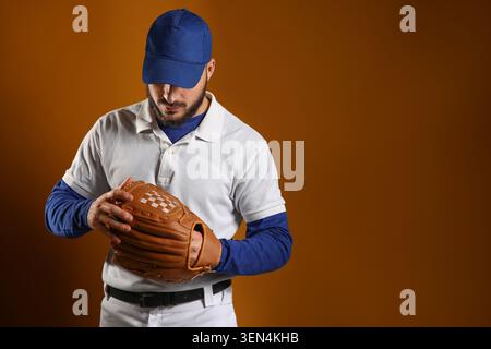 Baseball player in blue cap and white polo shirt holding brown leather glove against orange background, the athlete is focused on his equipment, demon Stock Photo