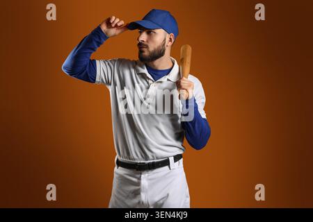 Young male baseball player in white uniform and blue cap holding bat, sports and athletic lifestyle, he stands confidently against an orange studio ba Stock Photo