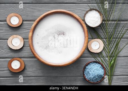 Spa relaxation with wooden bowl of soapy water, candles, coconut cream, and blue sea salt on gray wood Stock Photo
