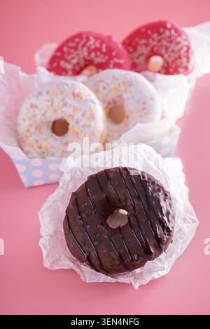 Assorted glazed donuts with sprinkles and chocolate drizzle on pink background, sweet bakery treats, these delicious pastries are individually wrapped Stock Photo